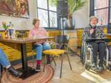Vrouw zit met map op schoot en microfoon in hand met aan de linkerkant publiek.