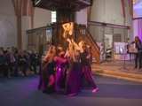 Dansgroep op podium met microfoon in Nicolaikerk bij aftrap 80 jaar vrijheid. In de achtergrond word viool gespeeld.